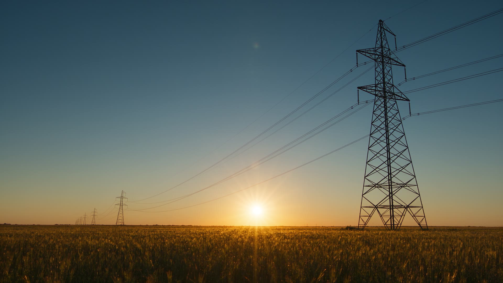 A high-voltage transmission tower and power lines over an open field at sunrise